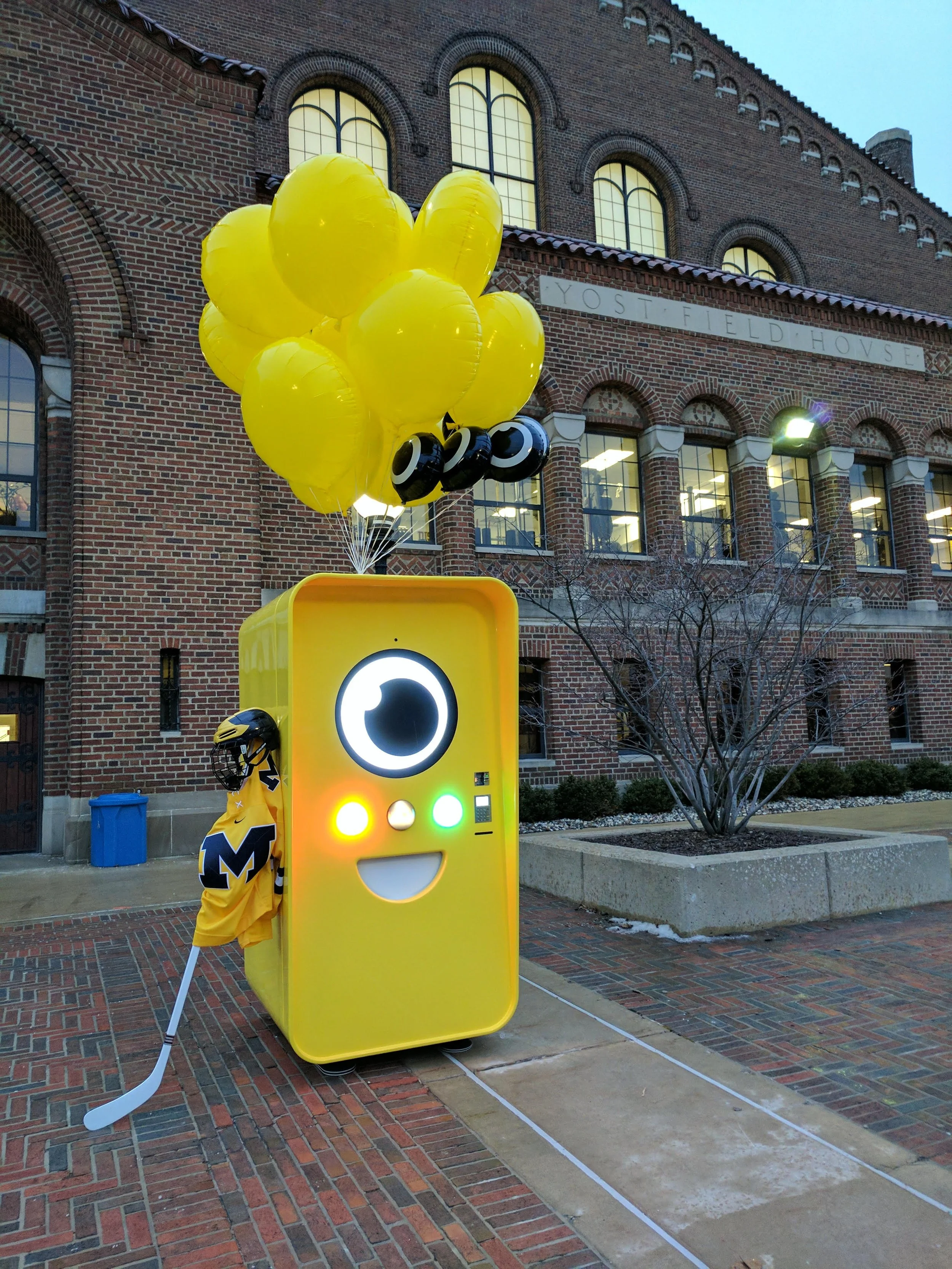 Yellow Snapbot vending machine with balloons outside Yost Field House, a Michigan hockey jersey draped beside it